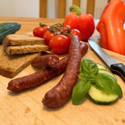Auf einem Holzbrett liegen Wurst, Tomaten, Gurke, Paprika und Brot.