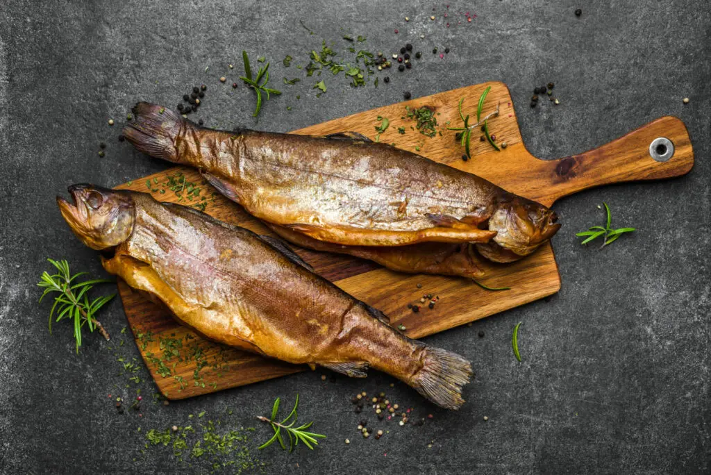 Zwei geräucherte Fische auf einem Holzbrett mit Kräutern und Gewürzen.