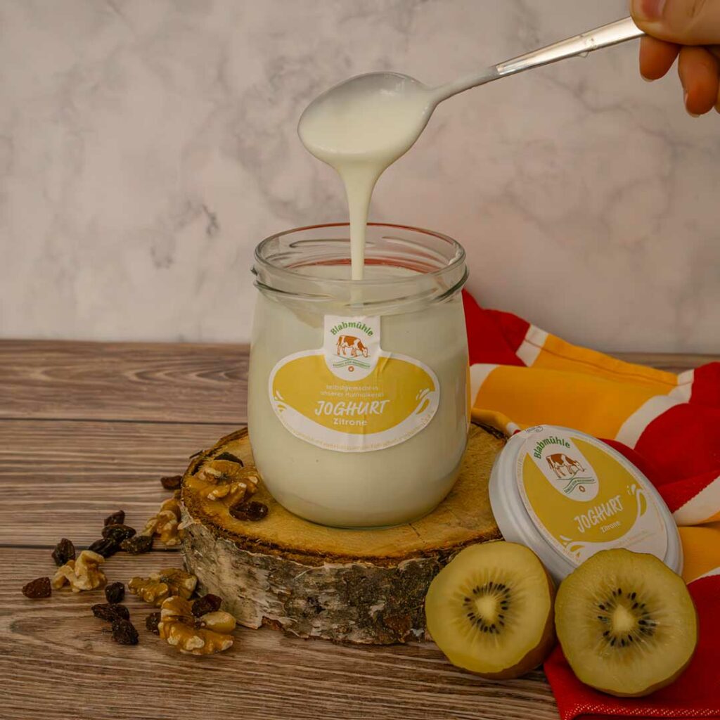 Ein Glas Zitronenjoghurt mit einem Löffel, der Joghurt herausnimmt, auf einem Holzbrett.