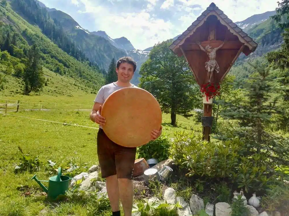 Mann in traditioneller Kleidung hält einen großen runden Käselaib in den Alpen. Im Hintergrund eine Berglandschaft mit grünem Tal, Bäumen und einem hölzernen Wegkreuz mit Kruzifix.