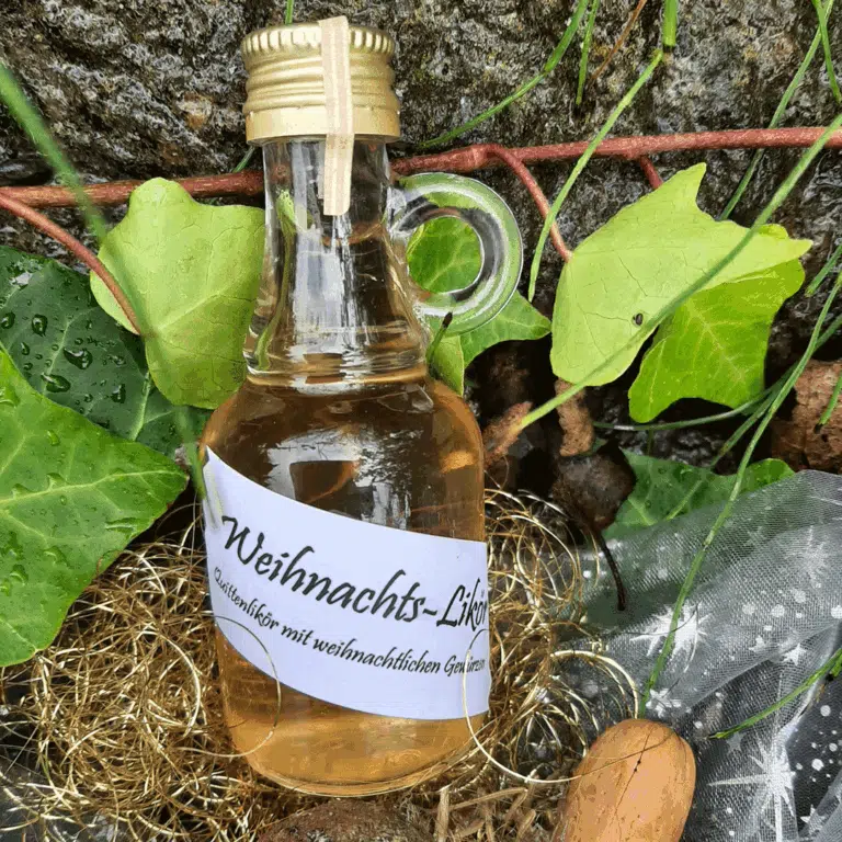 Kleine Glasflasche mit goldfarbenem Schraubverschluss und Henkel, gefüllt mit hellem Weihnachtslikör. Die Flasche trägt ein Etikett mit der Aufschrift ‚Weihnachts-Likör – Likör mit weihnachtlichen Gewürzen‘. Arrangiert inmitten von grünem Efeu, Naturmaterialien und goldener Deko.