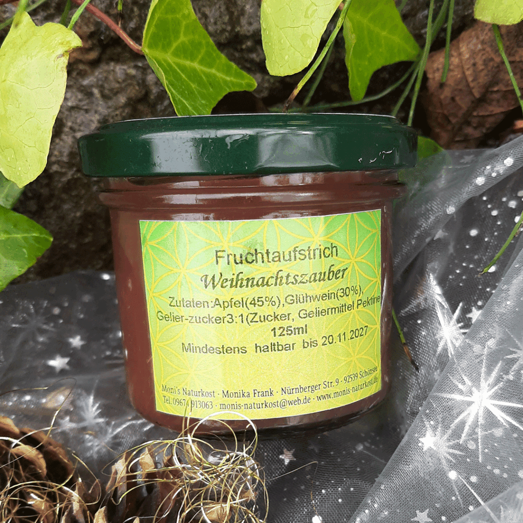 Glas mit dunkelgrünem Schraubdeckel, gefüllt mit Fruchtaufstrich ‚Weihnachtszauber‘. Das gelb-grüne Etikett nennt die Zutaten: Apfel (45 %), Glühwein (30 %), Gelierzucker 3:1. Das Glas steht dekorativ zwischen Efeublättern und weihnachtlicher Deko.