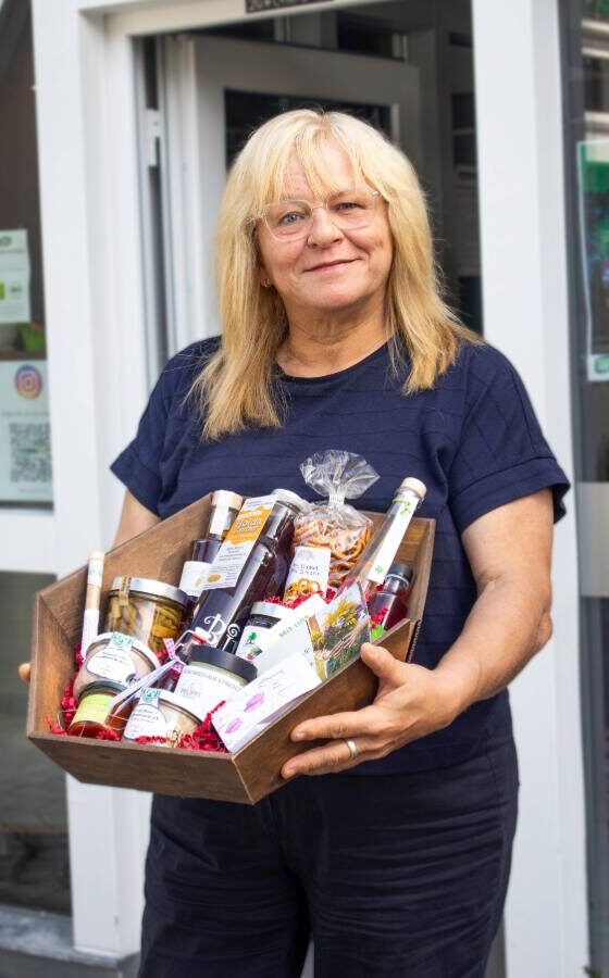 Frau mit blonden Haaren und Brille hält einen Holzkorb voller regionaler Produkte und Feinkost vor einem Ladeneingang.