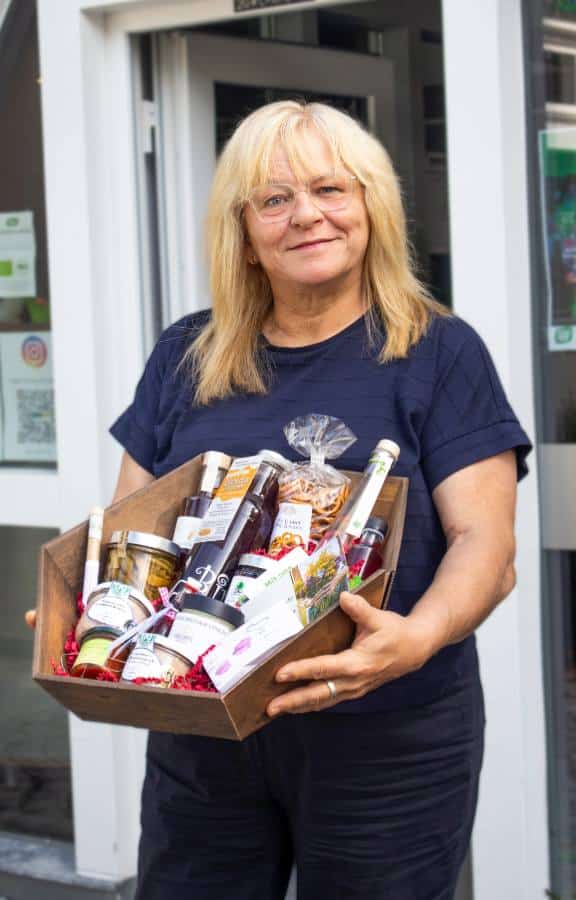 Frau mit blonden Haaren und Brille hält einen Holzkorb voller regionaler Produkte und Feinkost vor einem Ladeneingang.