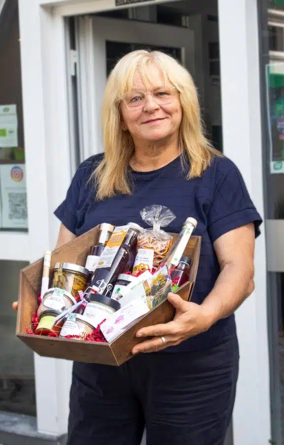 Frau mit blonden Haaren und Brille hält einen Holzkorb voller regionaler Produkte und Feinkost vor einem Ladeneingang.